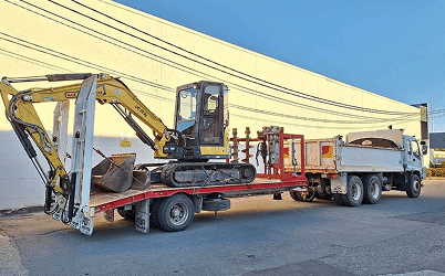 Flatbed lorry carrying a small yellow digger and earthmoving equipment, parked by a large building in Brisbane.